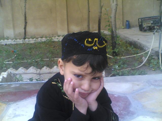 A young boy in an embroidered black vest and cap, resting his chin on his hands in a garden